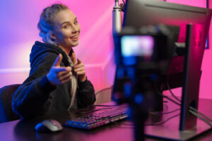 Funny and smiling e-sport gamer woman streaming live vlog while playing online video game on her PC. Room with blue and pink colored LED lights.